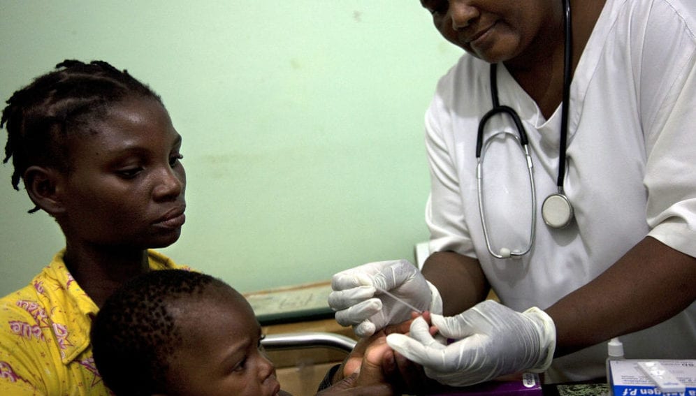 A woman sits with her child as a health worker takes a finger prick blood sample