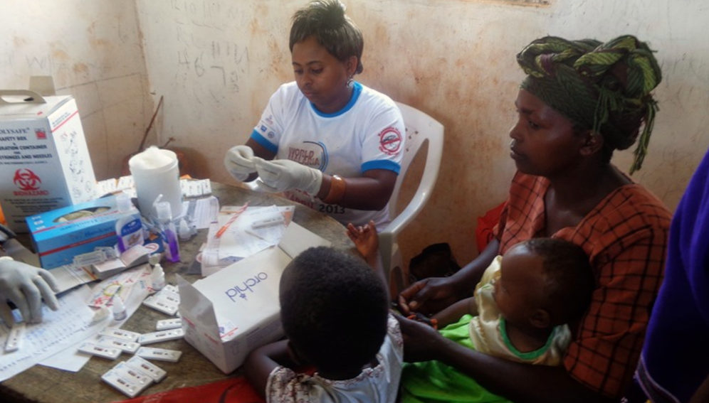 A health worker performs rapid diagnostic test (RDT) on a mother and her children. Source: Wikimedia.