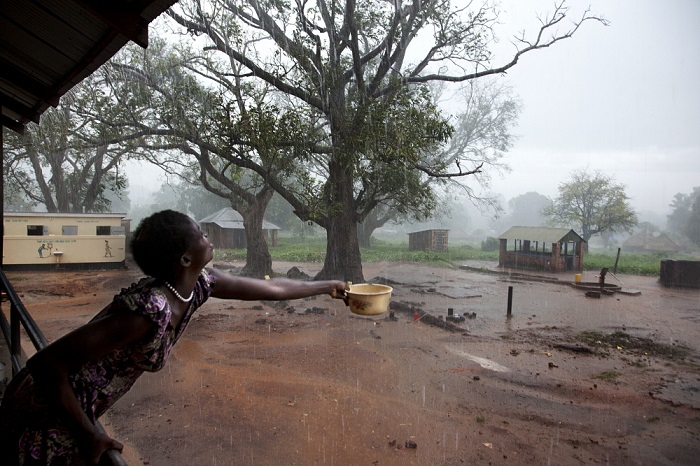woman standing on the verandah of a building holds out a bowl to collect rainwater